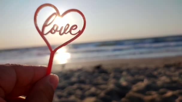 晴れた夏の朝に海の波の近くの海岸の砂浜で輝く朝の太陽の背景にハートとワードラブの形で手の赤い棒を保持する女性 ロマンチック セント バレンタインデー — ストック動画