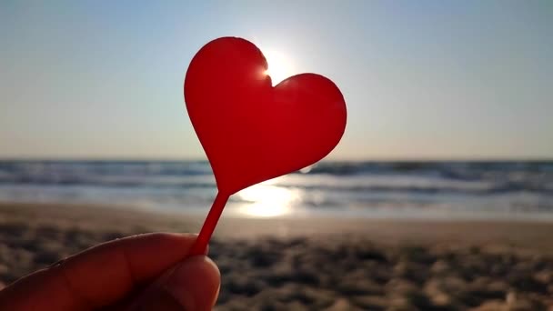 晴れた夏の朝に海の波の近くの海岸の砂浜に輝く朝の太陽の背景にハートの形をした手の赤い棒を握る女性 ロマンチック アモラス セント バレンタインデー — ストック動画