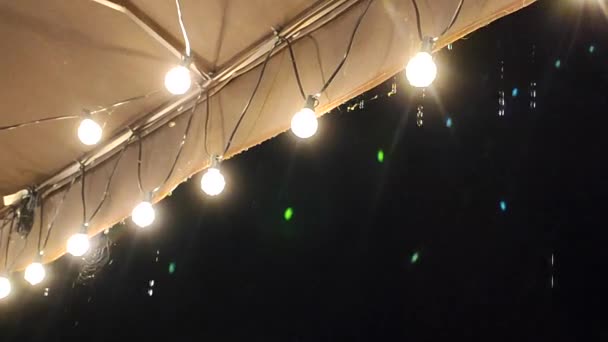 빛나는 등불이있는 지붕에서 떨어지는 어두운 저녁에 전구의 화환으로 덮개의 가장자리에서 — 비디오