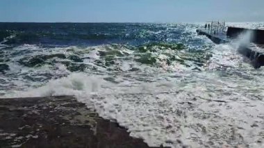 Fırtınalı deniz. Büyük deniz dalgaları güneşli yaz gününde eski beton iskeleye çarpıp çarpıyor. Beyaz köpüklü yüksek dalgalar. Büyük dalgalar kopuyor, çimento iskelesine yayılıyor. Patlamalar ve sıçramalar.