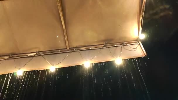 雨点般的雨点般从屋顶上滴下 夜晚灯火通明 黄昏时分 从雨篷边缘喷出的细雨带着灯泡的花环 大雨倾盆和雨滴 — 图库视频影像