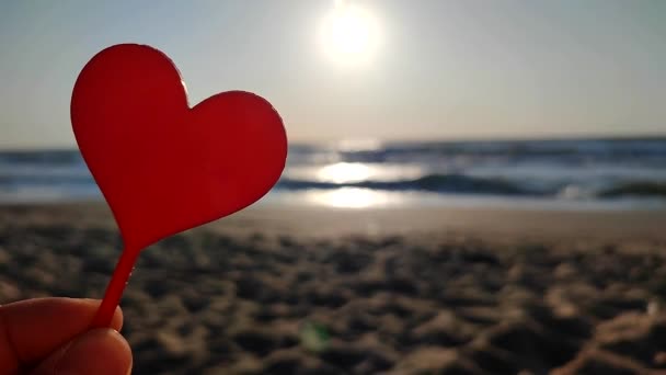 晴れた夏の朝に海の波の近くの海岸の砂浜に輝く朝の太陽の背景にハートの形をした手の赤い棒を握る女性 ロマンチック アモラス セント バレンタインデー — ストック動画