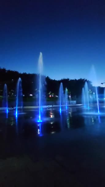 夜のカラフルなライトで照らされた歩行者地の噴水 市内の多彩な光に照らされた大きな公共の街の噴水 水のジェットが上昇して落ちる ウォーター — ストック動画