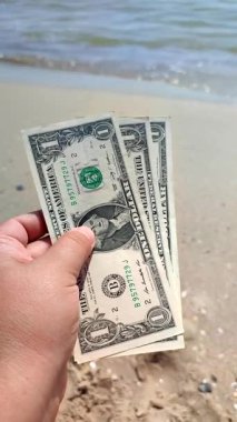 Elinde, kumlu sahildeki deniz dalgalarının arka planında bir dolar değerinde üç banknot tutan kişi. Güneşli bir yaz gününde deniz kıyısında. Konsept para finans Dikey