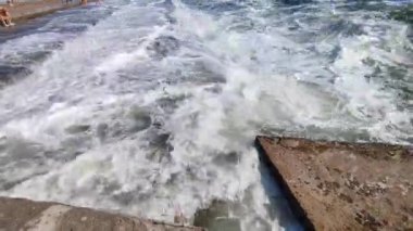 Birçok insanın şehirde durduğu eski çimento kıyısına büyük bir dalga çarpıyor. Güneşli bir günde dalgalar iskele iskelesine vuruyor. Fırtına, fırtınalı deniz. Hava durumu. Beyaz köpüklü yüksek dalgalar.