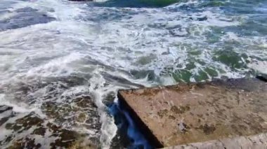 Birçok insanın şehirde durduğu eski çimento kıyısına büyük bir dalga çarpıyor. Güneşli bir günde dalgalar iskele iskelesine vuruyor. Fırtına, fırtınalı deniz. Hava durumu. Beyaz köpüklü yüksek dalgalar.