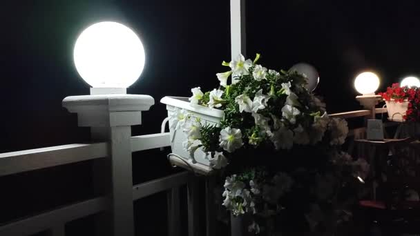 灯笼在漆黑的夜晚闪烁着光芒 白色的花朵在白锅中绽放 在风中摇曳 挂在靠近餐桌的栅栏上 晚上在咖啡馆里浪漫的气氛 户外装修室内装饰细节 — 图库视频影像