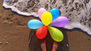 Deniz kıyısındaki ıslak kumsalda bir sürü renkli balon ve bir insanın ayaklarının dibinde dalgalanan dalgalar. Ağır çekim. Eğlence eğlencesi, dinlenme, eğlence, bayram kutlamaları.