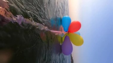 Güneşli bir sabah kıyı şeridi yakınlarında, su yüzeyinde kurdelelerle bağlanmış renkli küçük balonları çeken kişi. Yavaş çekim Dikey