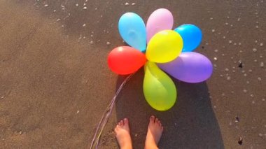 Deniz kıyısındaki ıslak kumsalda bir sürü renkli balon ve bir insanın ayaklarının dibinde dalgalanan dalgalar. Ağır çekim. Eğlence eğlencesi, dinlenme, eğlence, bayram kutlamaları.