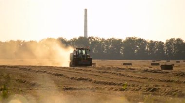 Güneşli bir yaz akşamında hasat makinası samanı balya sürüş sahasına sıkıştırıyor. Buğday balyalarıyla dolu bir tarla. Sahada çok toz var. Tarım endüstrisi tarımsal hasat çalışmaları