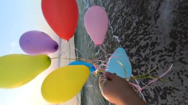 Gün doğumunda ve günbatımında deniz kıyısının kumlu sahilinde kurdeleyle bağlanmış bir sürü renkli balon ve el. Şafakta çok renkli balonlar