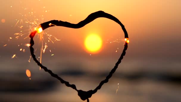 海の背景に燃えるハート型のスパークラー 夜明けに太陽が昇る ベンガル火災は 日の出の夜明けと海の日没に輝く心の形で インフレーション バレンタインデー — ストック動画
