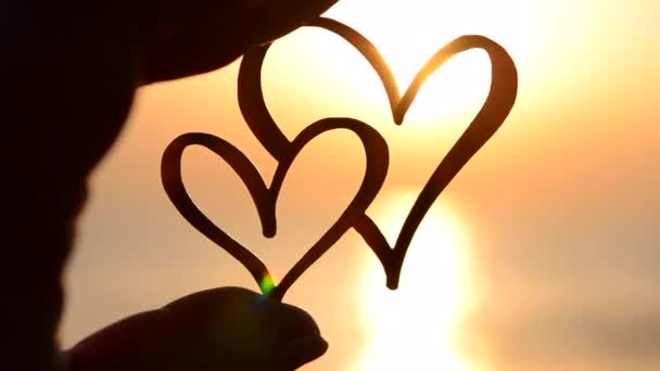 夜明けと海の日没に太陽の背景に対して2つの心の形をした黒い輪郭を指で保持する女性 コンセプト愛 ロマンチックなバレンタインデー気分の感情 — ストック動画