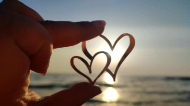 İki kalp şeklinde parmakları olan bir kadın şafak vakti güneşin arka planında ve denizde gün batımında iki kalp şeklinde duruyor. Romantik sevgililer günü duygusal ilişki kavramını kavra.