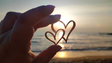 İki kalp şeklinde parmakları olan bir kadın şafak vakti güneşin arka planında ve denizde gün batımında iki kalp şeklinde duruyor. Romantik sevgililer günü duygusal ilişki kavramını kavra.