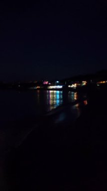Deniz kıyısındaki binaların parlayan ışıklarının yansıması. Gece deniz dalgalarının sularında. Deniz kıyısında ışıklı lambaları olan binalar. Gece deniz kıyısındaki şehir. Suda aydınlanma. Dikey