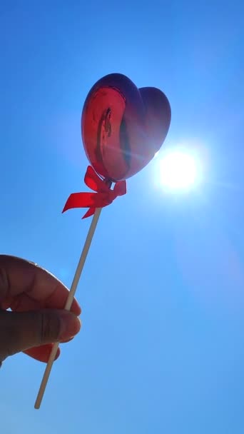 女性の体積の赤い心臓の形をした棒は 青い空の背景と晴れた夏の日に輝く太陽に対して手を差し伸べます コンセプトの愛 バレンタインデー 恋に落ちる バーティカル — ストック動画