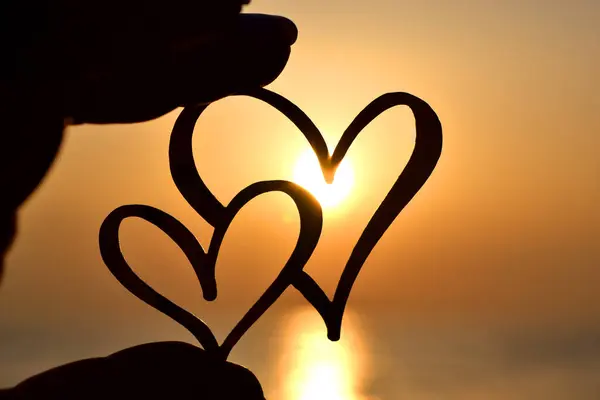 Woman holding in fingers black outline in shape of two hearts against background of sun at dawn and sunset on sea. Concept love infatuation romantic Valentines Day mood emotions relationship