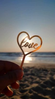 Parmaklarını tutan kişi, altın kalp şeklinde altın bir kalbe ve arka planda aşk kelimesine sarılır. Güneşli yaz günlerinde deniz dalgaları yakın planda olur. Aşk romantizmi kavramı Aziz Sevgililer Günü Dikey