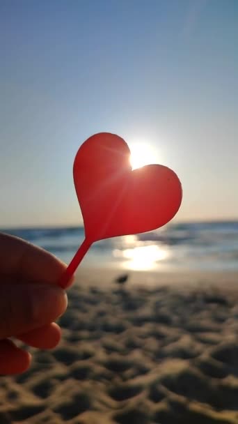 バックグラウンドの海と海の波 晴れた夏の日の終わりの海岸の青空の形の赤い心臓に指の手で握っている人 コンセプト愛ロマンスアワーセントバレンタインデー垂直 — ストック動画