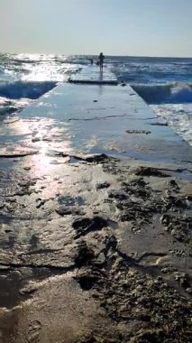 Islak elbiseli genç bir kadın güneşli bir yaz gününde ıslak bir iskele boyunca insanlara doğru yürüyor. Dalgalar iskeleye çarpıyor. Bir sürü büyük sıçrama. Güneşli bir yaz gününde düşey denizde fırtına