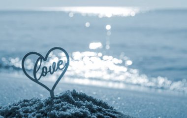 Plastik çubuk kırmızı kalp şeklinde ve kelime aşk kumsalda kumsalda güneşli yaz günlerinde yakın plan. Kalp şeklinde figürler, deniz dalgalarında aşk. Aşk ilişkisi romantizmi. Mavi renk