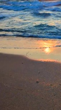 Deniz dalgaları güneşli bir günde şafak vakti kumlu sahil sahillerinde dalgalanır. Deniz dalgaları, köpükler ve kıyıya vuran kumlar. Sahil Kıyısı Sahili. Doğal arka plan. Doğa arkaplanı Yavaş çekim düşey