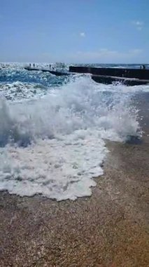 Fırtınalı deniz. Büyük deniz dalgaları güneşli yaz gününde eski beton iskeleye çarpıp çarpıyor. Yüksek dalgalar, beyaz köpük. Büyük dalgalar kopuyor, çimento iskelesine yayılıyor. Su sıçramaları dikey sıçrıyor