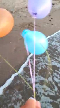 Güçlü rüzgarlarda denizin arka planında üç renkli şişme top. Elinde çok renkli hava balonlarıyla kumlu sahil kıyılarında arka planda deniz dalgalarıyla oynayan biri. Eğlence eğlencesi Dikey