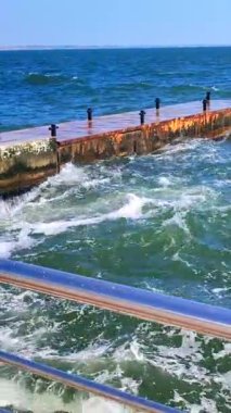 Büyük dalgalar, güneşli bir günde bir sürü sıçramayla ahşap iskeleye çarpıyor. Fırtına, fırtınalı deniz. Hava durumu. Seyahat, turizm, tatil. Sahil, sahil, sahil. Dikey