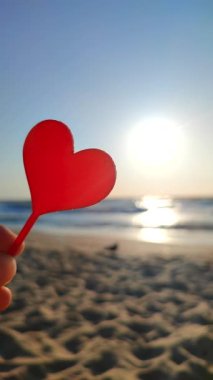 Parmaklarını tutan kişi arka planda kırmızı kalp, deniz ve deniz dalgaları, güneşli yaz günlerinde deniz kenarında mavi gökyüzü. Aşk romantizmi kavramı Aziz Sevgililer Günü Dikey