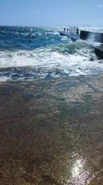 Fırtınalı deniz. Büyük deniz dalgaları güneşli yaz gününde eski beton iskeleye çarpıp çarpıyor. Yüksek dalgalar, beyaz köpük. Büyük dalgalar kopuyor, çimento iskelesine yayılıyor. Su sıçramaları dikey sıçrıyor