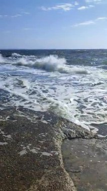 Fırtına. Denizde fırtına. Beyaz köpüklü büyük açık deniz dalgaları beton iskele duvarına çarpıyor ve güneşli bir yaz gününde su sıçrıyor. Fırtınalı deniz havası. Dalgalı deniz. Kıyıya. Doğa. Doğal arka plan. Dikey