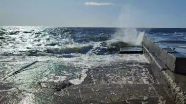 Fırtına. Denizde fırtına. Beyaz köpüklü büyük açık deniz dalgaları beton iskele duvarına çarpıyor ve güneşli bir yaz gününde su sıçrıyor. Fırtınalı deniz havası. Dalgalı deniz. Kıyıya ve dalgalara. Doğa. Doğal arkaplan