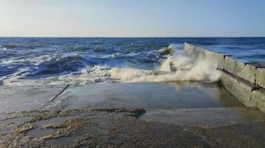 Fırtına. Denizde fırtına. Beyaz köpüklü büyük açık deniz dalgaları beton iskele duvarına çarpıyor ve güneşli bir yaz gününde su sıçrıyor. Fırtınalı deniz havası. Dalgalı deniz. Kıyıya ve dalgalara. Doğa. Doğal arkaplan