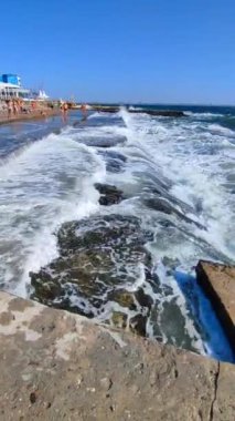 Beyaz köpüklü büyük deniz dalgaları, güneşli bir yaz gününde şehirde fırtına sırasında insanların üzerinde durduğu beton iskeleye çarpıp sıçrıyor. Fırtınalı deniz. Deniz manzarası, fırtına sırasında açık deniz dalgaları. Dikey