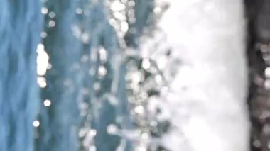 Deniz dalgaları yuvarlanıyor ve kayalara çarpıyor. Suyun yüzeyinde bir sürü beyaz güneş lekesi parlıyor. Güneşli yaz gününde deniz manzarası. Bulanık soyut doğal arkaplan dikey