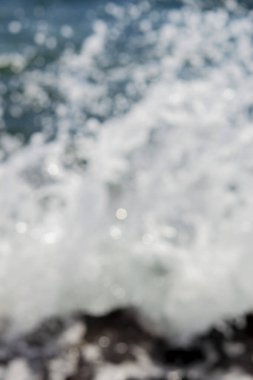 Deniz dalgaları yuvarlanıyor ve kayalara çarpıyor. Suyun yüzeyinde bir sürü beyaz güneş lekesi parlıyor. Güneşli bir yaz gününde deniz manzarası. Bulanık soyut doğal arkaplan