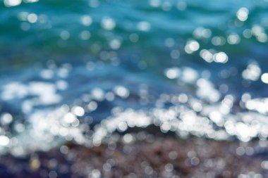 Deniz dalgaları yuvarlanıyor ve kayalara çarpıyor. Suyun yüzeyinde bir sürü beyaz güneş lekesi parlıyor. Güneşli bir yaz gününde deniz manzarası. Bulanık soyut doğal arkaplan