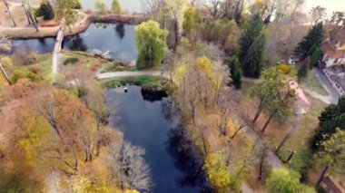 Kirli yolları, köprüleri ve bir sonbahar günü yürüyen insanlarla dolu bir parktaki mavi su göllerinin üzerinde uçmak. Bir sonbahar günü kırmızı, sarı ve yeşil yapraklı birçok ağaç ve çalı. Hava aracı görünümü