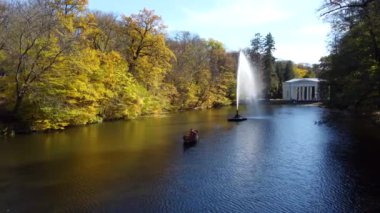 Beautiful landscape people sailing in boat on lake with decorative fountain among trees with yellow leaves in park on sunny autumn day. Natural background. Travel tourism rest recreation. Aerial view