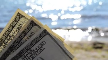 Güneşli bir günde denizin arka planında dolar banknotları ve deniz kenarında kumlu kumsalda güneşli bir yaz günü yakın plan. 100 dolarlık banknot gömülü. Para Amerikan 100 dolarlık banknotlar kum ve denizin yanında. Rüzgarlı. Finans
