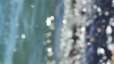 Deniz dalgaları yuvarlanıyor ve kayalara çarpıyor. Suyun yüzeyinde bir sürü beyaz güneş lekesi parlıyor. Güneşli yaz gününde deniz manzarası. Bulanık soyut doğal arkaplan dikey