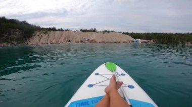 Taş ocağı gölünün mavi sularında yüzen bir SUP tahtasındaki kız. Üzerinde kayalık kıyılar ve ağaçlar olan bir yaz günü. Yaz sporları, turizm, dinlenme, rahatlama, seyahat, dinlenme