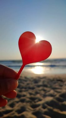 Parmaklarını tutan kişi arka planda kırmızı kalp, deniz ve deniz dalgaları, güneşli yaz günlerinde deniz kenarında mavi gökyüzü. Aşk romantizmi kavramı Aziz Sevgililer Günü Dikey