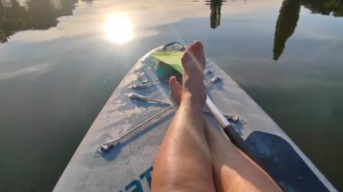 Nehir ya da gölün yüzeyinde çıplak kadın bacakları sıcak yaz akşamını yansıtıyor. Sakin bir günde SUP panosunda yüzen biri. Turizm seyahati, dinlenme sporu yaşam tarzı etkinliği