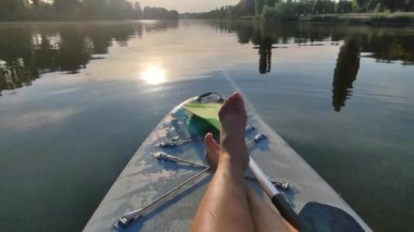 Nehir ya da gölün yüzeyinde çıplak kadın bacakları ve sıcak yaz akşamlarında da ağaçlar. Sakin bir günde SUP panosunda yüzen biri. Turizm sporu