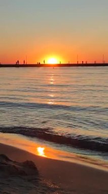 Denizde şafak vakti. İnsanlar güneşin doğuşuna karşı siyah bir iskelede yürüyorlar. Dalgalar deniz kıyısının kumlu sahillerine doğru yuvarlanıyor. Deniz suyunun yüzeyindeki güneşli yol. Sakin rüzgarsız hava. Dikey