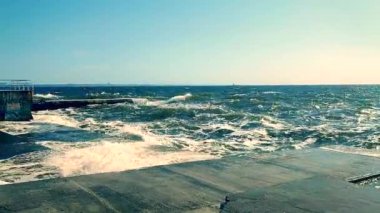 Güneşli yaz gününde denizde fırtına. Güneşli bir günde beton iskeleye beyaz köpüklü büyük deniz dalgaları çarpıyor. Büyük dalgalar çimento iskelesine ve temiz mavi gökyüzüne çarpıyor.